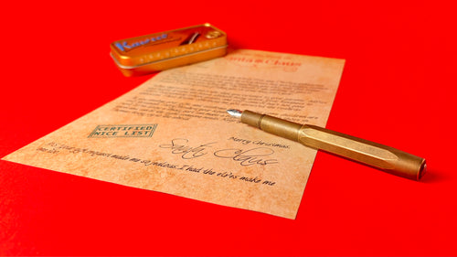 A gold-toned metal fountain pen with a clear glass barrel lies diagonally across a vintage-style parchment letter on a solid red background. A small gold metal tin case is visible in the upper left corner.