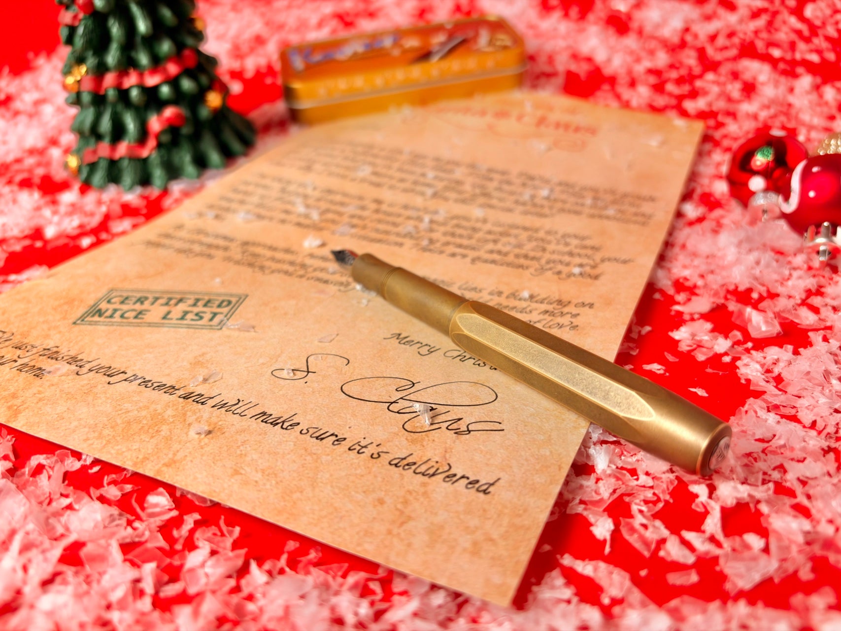 A gold metal pen resting on a parchment-style Christmas letter, with a small decorated tree and red festive background.