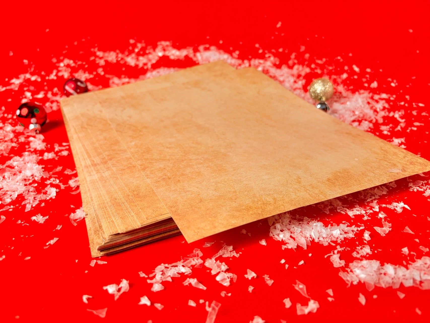 A stack of textured kraft paper sheets or cards arranged diagonally on a bright red background, surrounded by scattered white flakes and small decorative holiday ornaments.