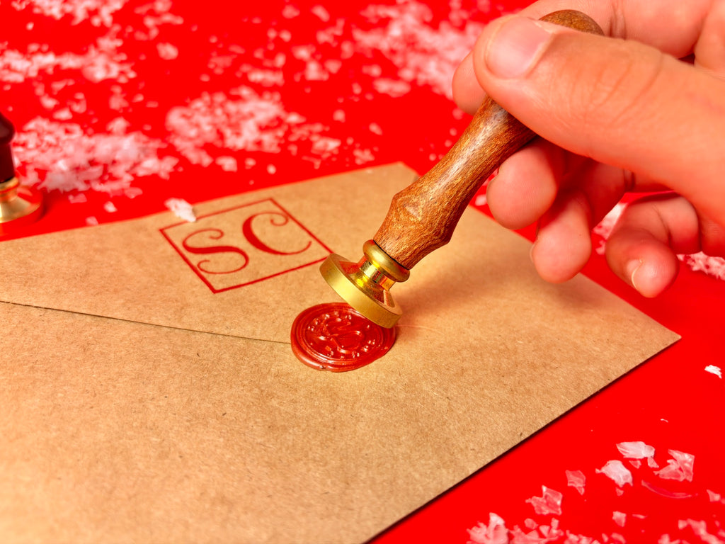 A hand holds a wooden-handled wax seal stamp with a gold-toned metal base, pressing it onto a brown envelope to seal it with red wax. The stamp leaves a red square stamp with the initials 'SC' on the envelope, set against a bright red background with scattered white flakes.