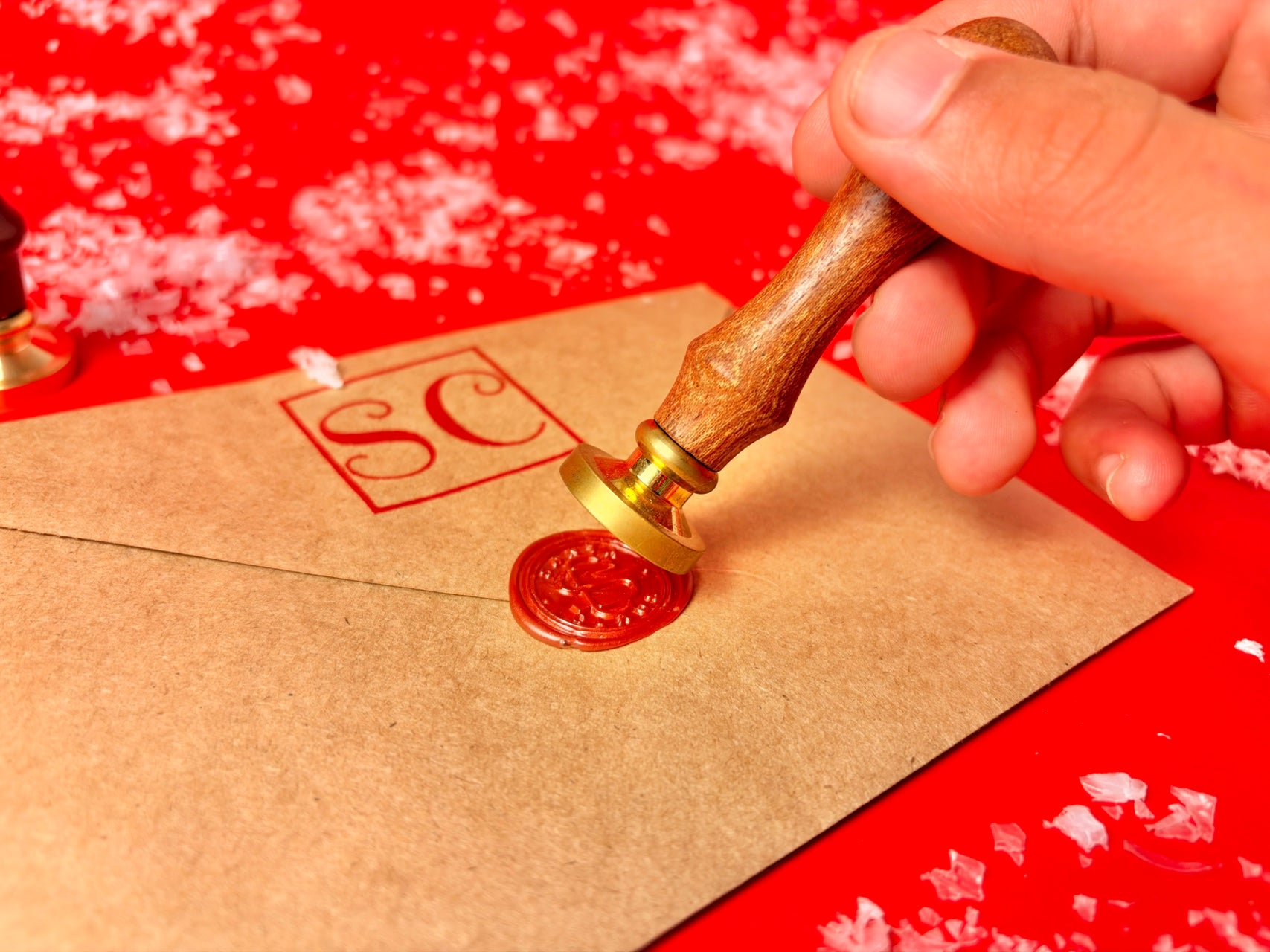 A hand holds a wooden-handled wax seal stamp with a gold-toned metal base, pressing it onto a brown envelope to seal it with red wax. The stamp leaves a red square stamp with the initials 'SC' on the envelope, set against a bright red background with scattered white flakes.