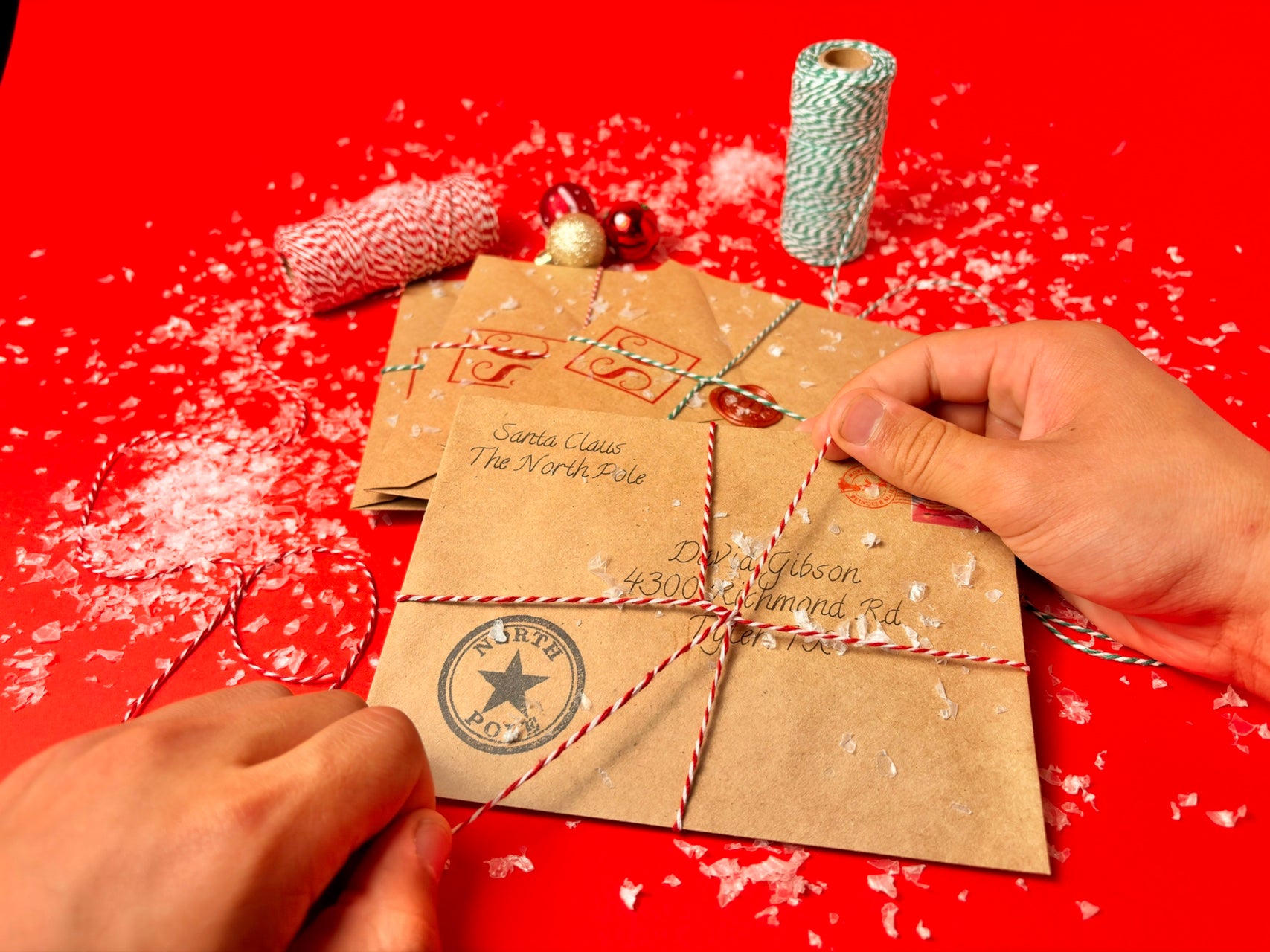 A close-up of hands wrapping kraft paper holiday envelopes with red-and-white striped twine on a bright red background, with additional twine spools and decorative elements nearby.